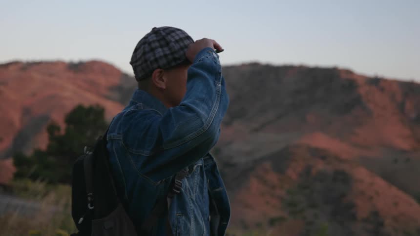 Man adjusts his hat while looking out over a hilly area at sunset