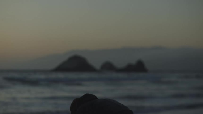 A woman on a beach looks out to the sea with her hands in her coat pocket