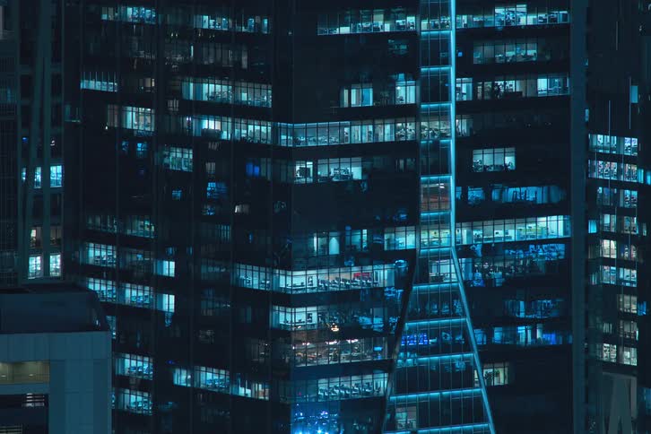 A glass building mirroring the city lights turn on at the same time at night