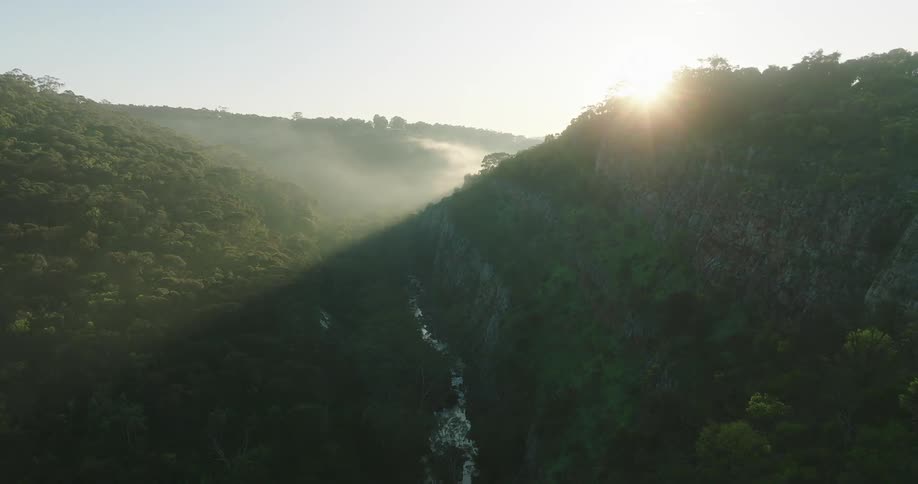Aerial view of a central river amidst mountainous terrain filled with vegetation with the sun on the horizon
