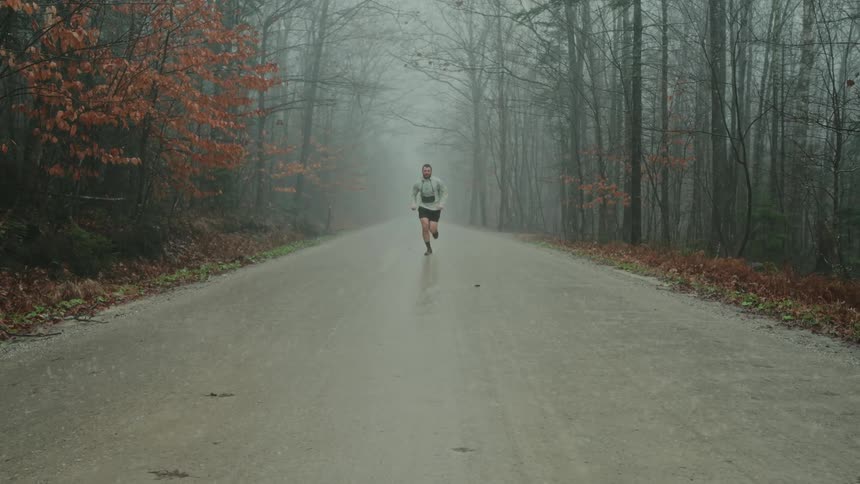 In the forest a bearded man in a gray hoodie and black shorts jogs along a dirt road in the pouring rain
