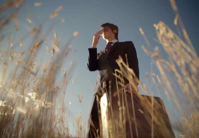 A businessman clad in a suit with sunglasses and carrying a briefcase stands in a field
