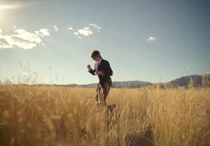 A man dressed in a suit and holding a suitcase stands on a grassy field under the sun he puts on a pair of sunglasses