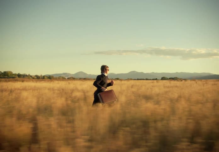 A man in a suit and briefcase is running in a field of tall vegetation and soon he falls