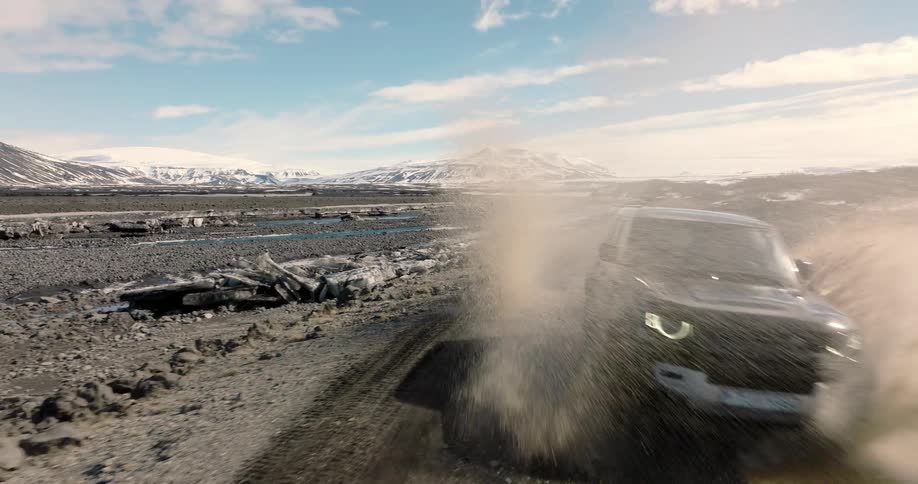 Car passing through puddles on a dirt road next to a river with a chain of mountains in the background covered in snow