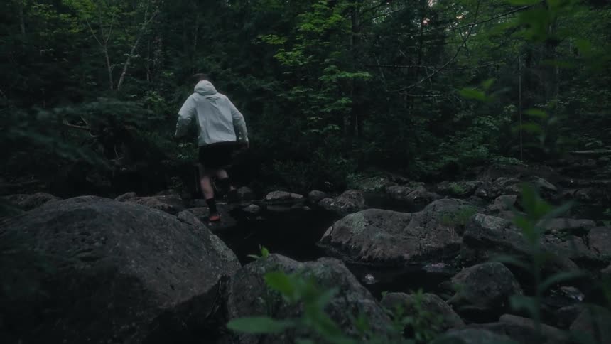 Man in a white hoodie jumps from one rock to another rock over a stream in the forest