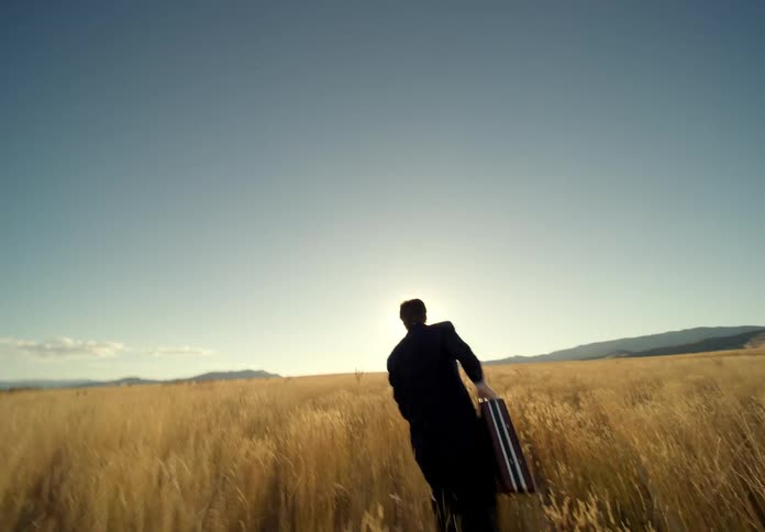 A man in a suit and carrying a briefcase runs across a field until he stops looking tired and scared