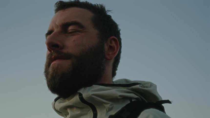 A bearded man wearing a hoodie and a backpack is gazing in the distance as he prepares for a hike