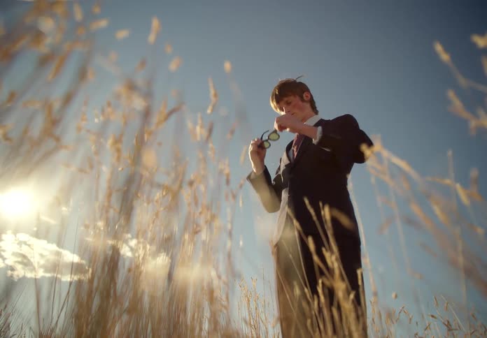 Man in a suit in the middle of a pasture taking off his glasses cleaning them and continuing to walk