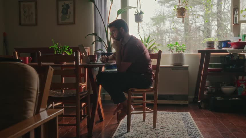Bearded man with a t shirt and pants sits at table using a laptop in the living room