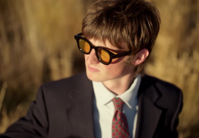 A man in a field puts on glasses and looks in the direction of the sun holding up his hand for protection