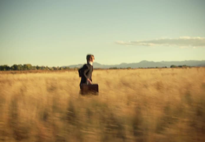 Man in a suit running through a pasture with a suitcase during the day