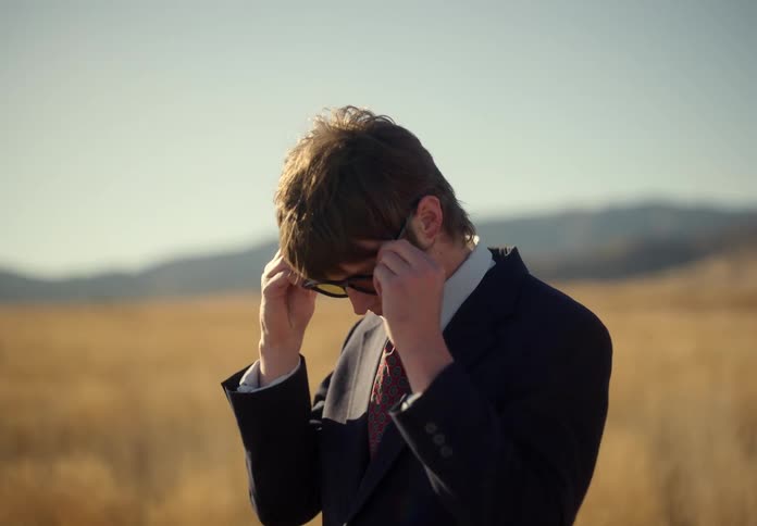 Man in a suit in a field cleaning his glasses looking up and walking