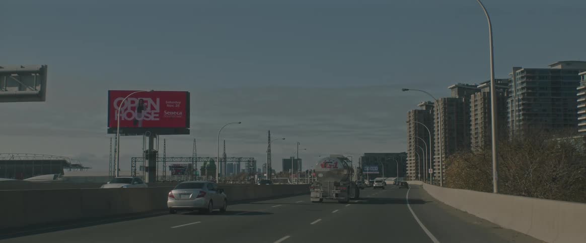 Cars and a fuel tanker truck drive down a highway in an urban area on a sunny day