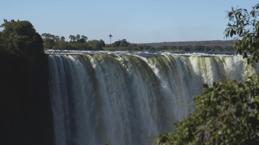 Waterfalls flow over a cliff with trees in the distance