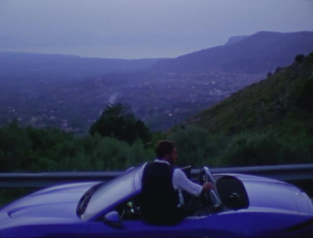 Man sitting on compact car parked on road way overlooking foggy town gestures