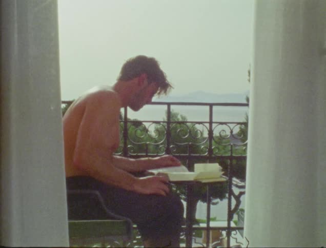 Shirtless man sitting on the balcony writing in a book