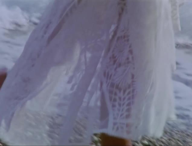A woman wearing sunglasses and a white transparent gown is walking on the beach on a sunny day