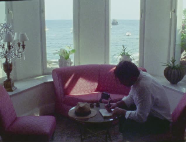 Man wearing a white shirt sitting down writing on a piece of paper on a coffee table indoors during day time