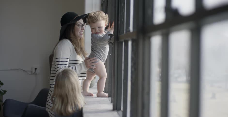A child walks up to a woman with a baby by a window