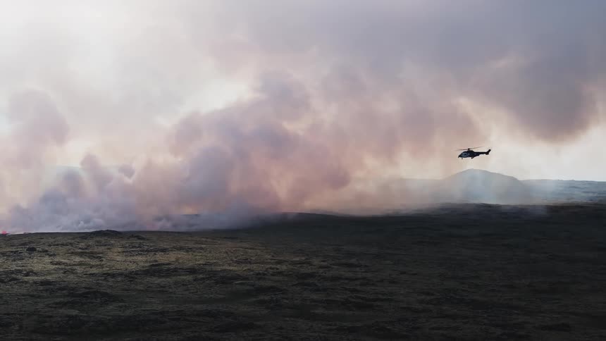 Helicopter flying over a field near a volcano with smoke rising