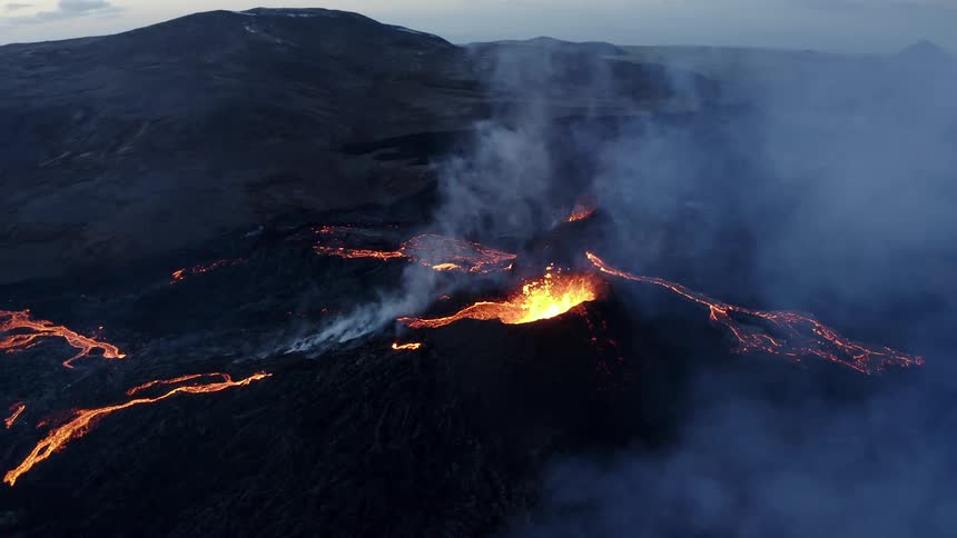 Molten hot lava glows as it splashes from a volcano and smoke rises as lava flows