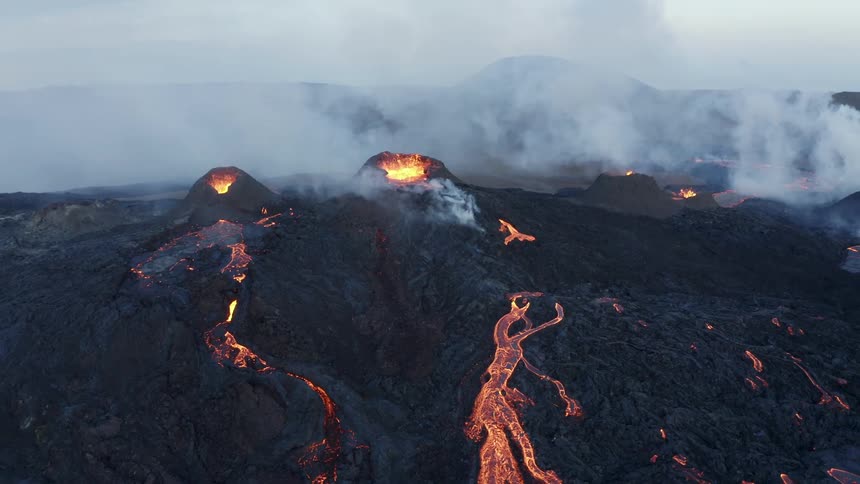 Aerial of smoke rising from multiple erupting volcanoes with lava flowing over ash covered ground