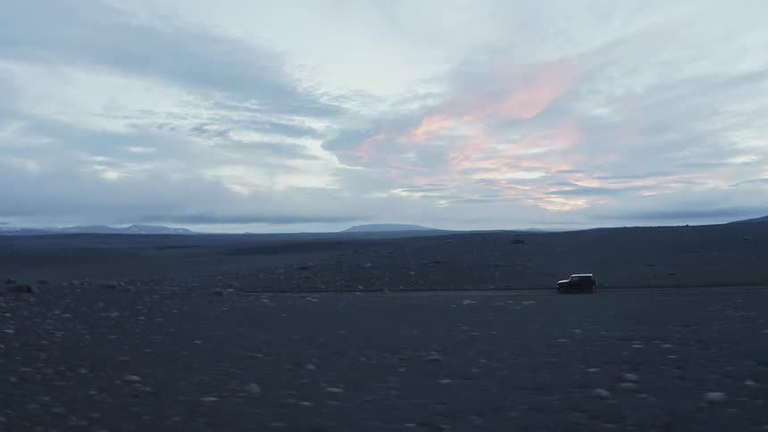 Vehicle drives along highway through bare earth rock strewn terrain under red tinted clouds