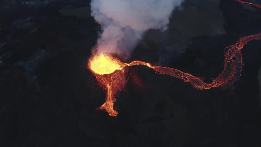Molten hot lava glows in the darkness as it splashes from an erupting volcano and smoke rises as lava flows