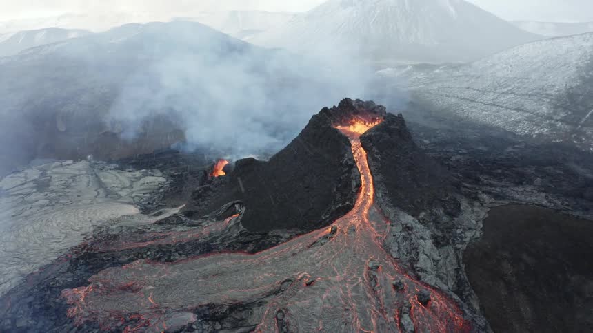 Molten hot lava glows as it splashes from an erupting volcano and flows across the land and snow flies among smoke in the air