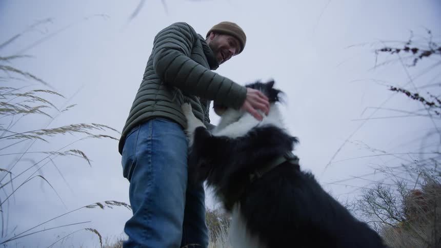Man wearing a coat and a beanie standing up outdoors playing with a border collie dog during day time