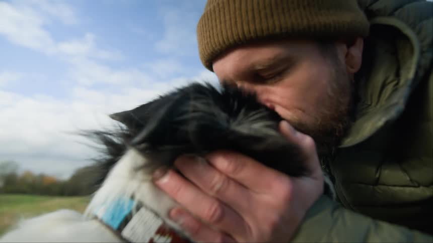 A man plays with and kisses his dog in a grassy field on an autumn day