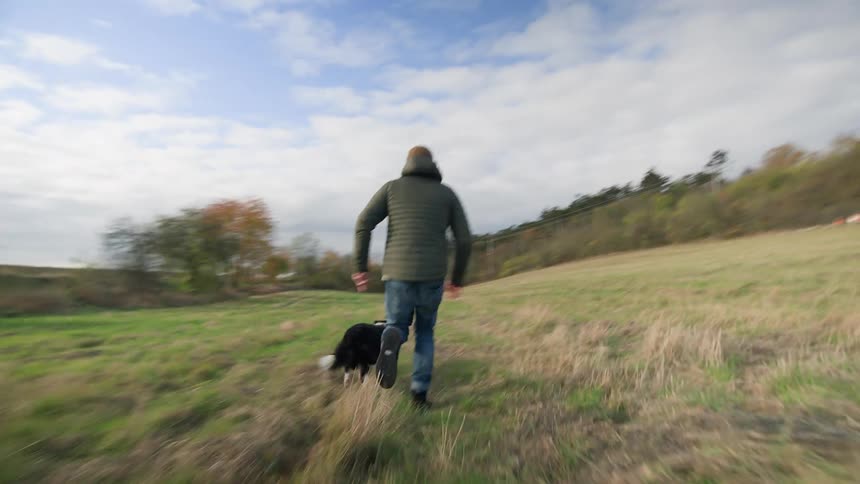 Man wearing a green jacket and jeans plays with dog in the green field