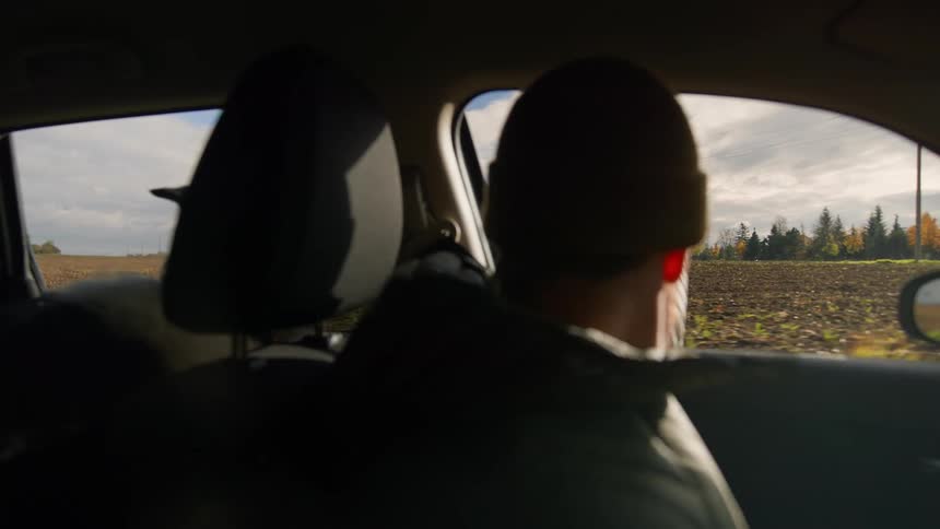Bearded man wearing a beanie driving a car on a road with a dog on the back seat during day time