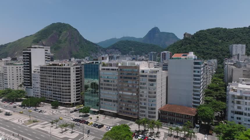 Aerial of a beach and hotels and buildings continuing back away from the coastline in between island hills with island mountains in the distance under a pale blue sky during the day
