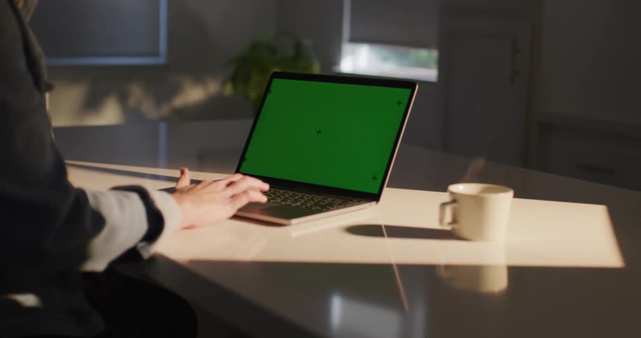 A man is sitting at a table typing on a laptop computer with a green screen as steam rises from a cup of coffee