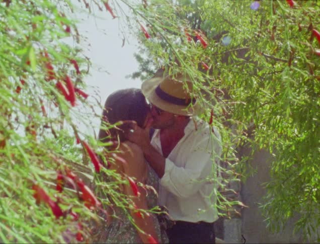 Man with hat sunglasses and white shirt makes out woman wearing sunglasses and sleeveless dress in garden next to trees