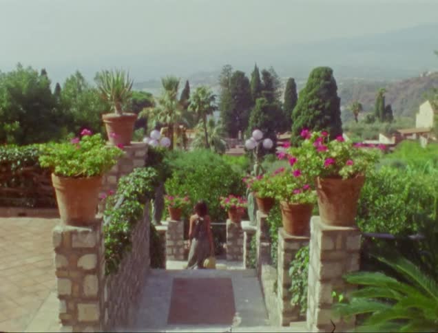 Man reads newspaper on terrace overlooking seashore while woman walks down steps through landscaping