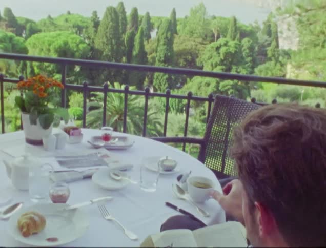 Man with beard and white shirt sits at table reading book and drinking coffee on balcony overlooking bay