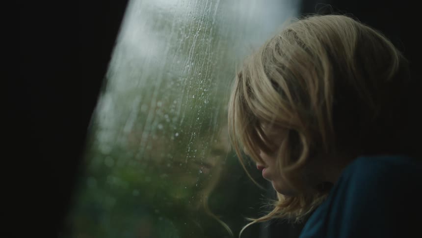 Boy rubbing his head against the window of the house while sadly watching the rain