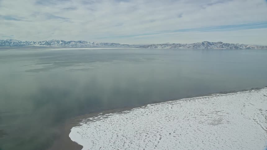 An aerial view of a sea next to mountains in a winter climate with snow on the ground and on top of the mountains