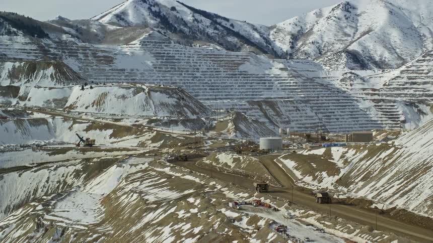 Aerial of dump trucks driving the road leading around to brigham canyon mine which is covered in a dusting of snow with snow covered mountain peaks in the background on a bright day