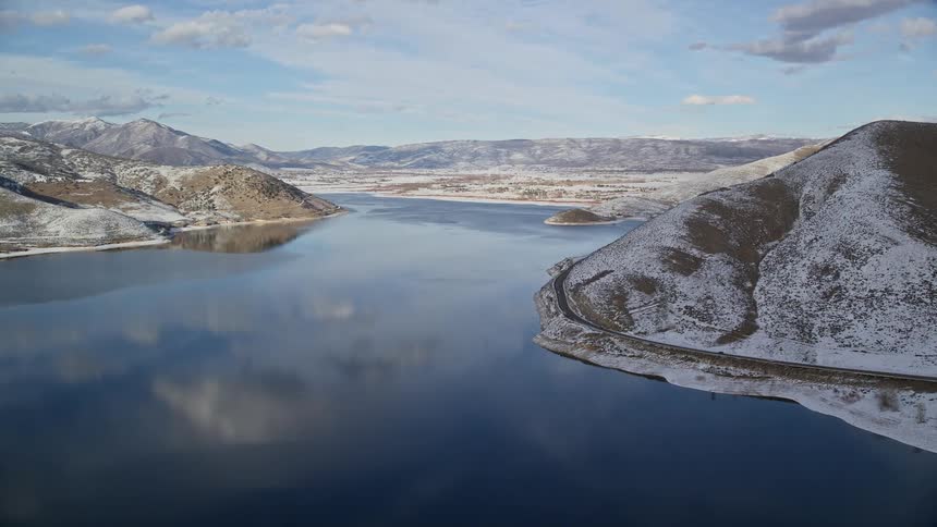 Aerial view of a lake with a curving shoreline next to a road on the right with moving cars surrounded by hills with a light snow cover reflecting clouds in the water