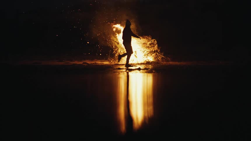A person walking around a campfire at the edge of a lake
