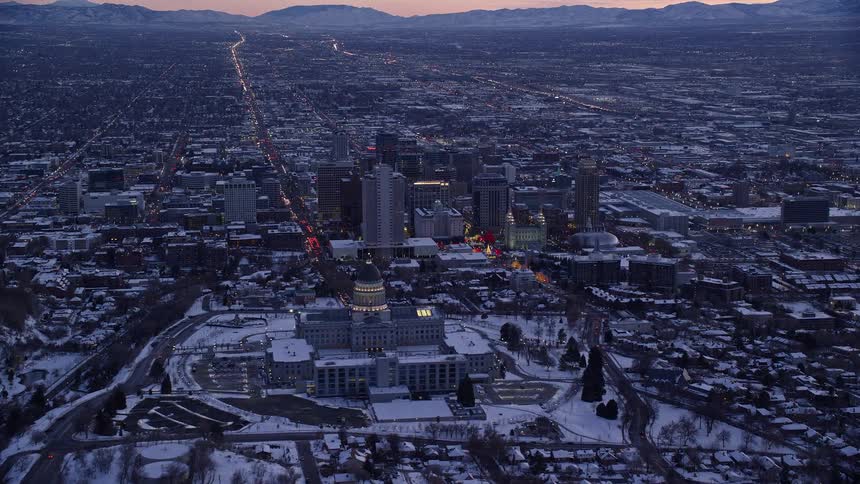 Aerial of salt lake city with a pink sky and purple mountain range in the background closing in on the capital building with snow on the ground at dusk
