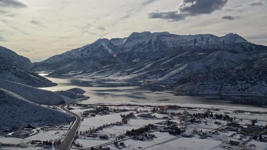 Vehicles drive along wide highway through lake side town bordering mountain on a winter day