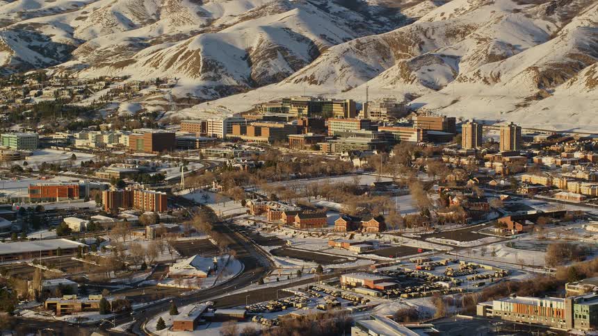 Aerial at the edge of the city with neutral colored buildings and snow on the ground beside neutral colored snow covered mountain peaks in the sunlight
