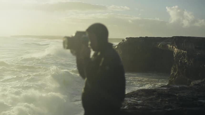 A cameraman taking photos of the sea and behind him violet waves are breaking on the rocks that are on the shore