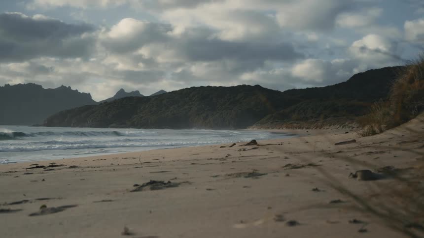 Waves roll into shore and over the beach with mountains along the coast under a cloudy sky
