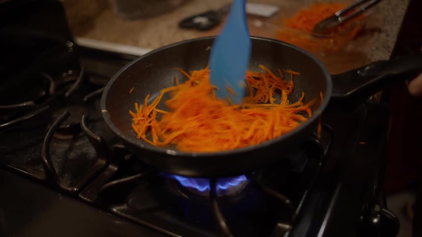 A person stirring some julienne carrots with a blue spatula in a pan on a gas stove top